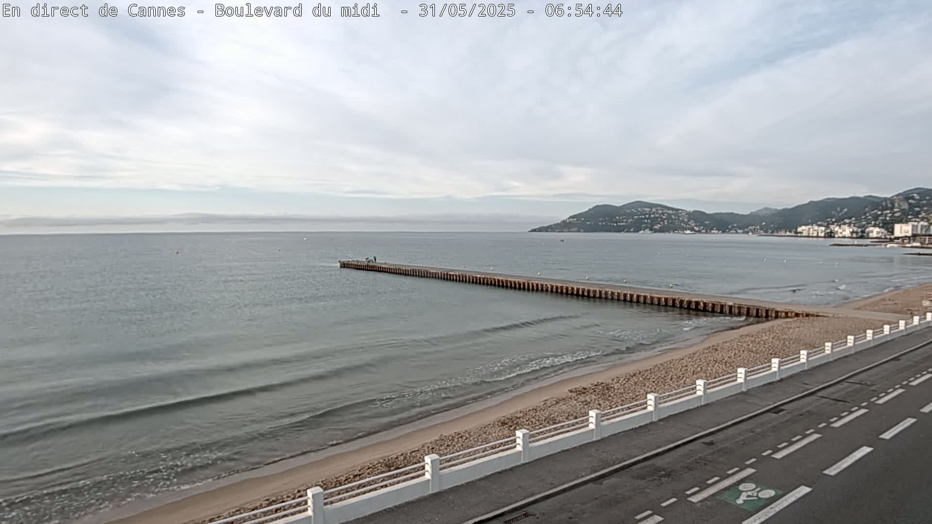 A long pier extends into a calm sea with small waves lapping a sandy beach, all under a partly cloudy sky.