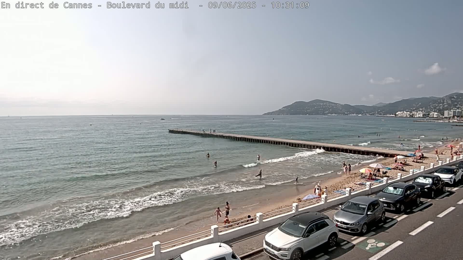 A sandy beach with people swimming and sunbathing stretches along a calm sea under a mostly clear sky, with cars parked along the roadside beside a low wall.