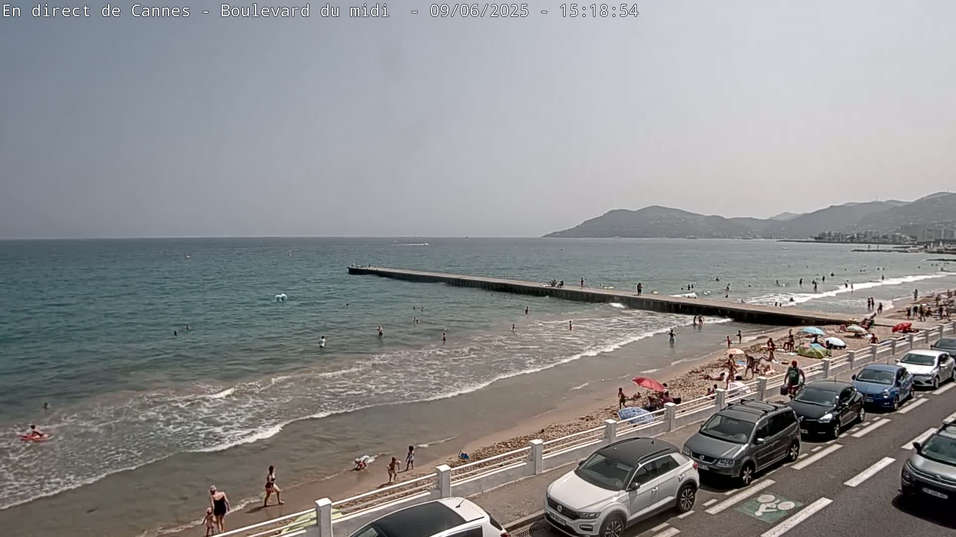 A sandy beach with many people swimming and sunbathing, a long pier extending into a calm sea under a hazy sky, and cars parked along a roadway next to the beach.