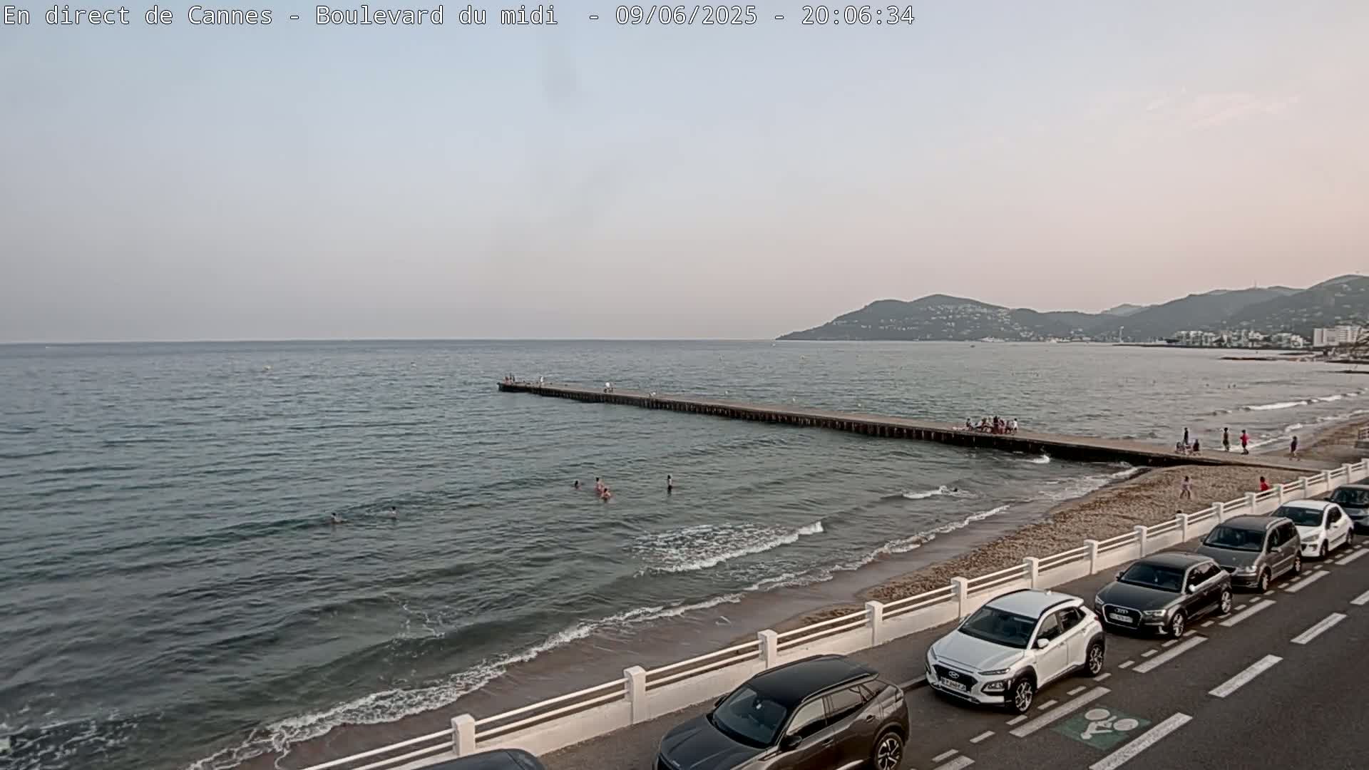 A long pier extends into a calm sea under a pale sky, with people swimming and walking along the shore where cars are parked.