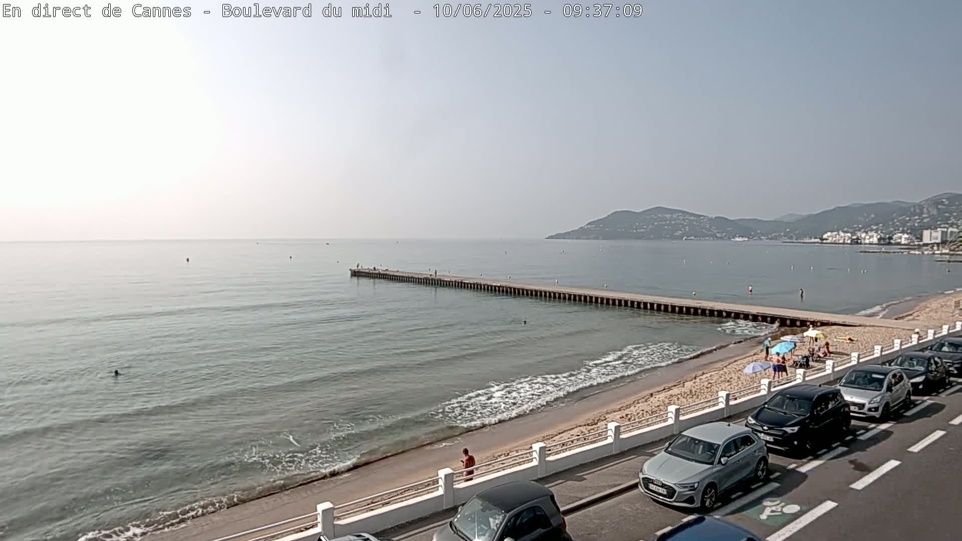 A sandy beach with calm ocean waters and a long pier extends into the sea under a hazy sky, with cars parked along a road overlooking the beach and a few people scattered on the sand and in the water.