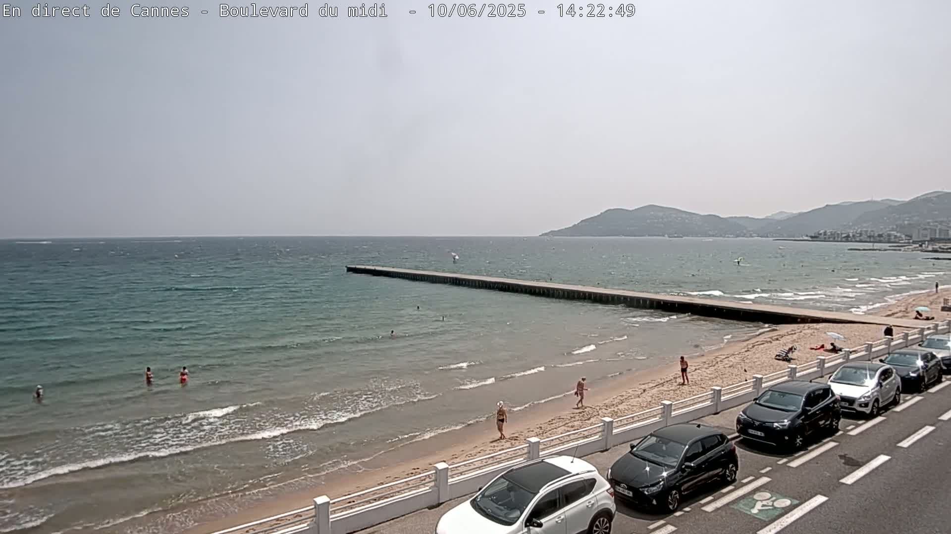 A sandy beach with calm, light blue water and several people swimming and sunbathing, a long pier extending into the sea, cars parked along a roadway bordering the beach, and distant mountains under a mostly clear sky.