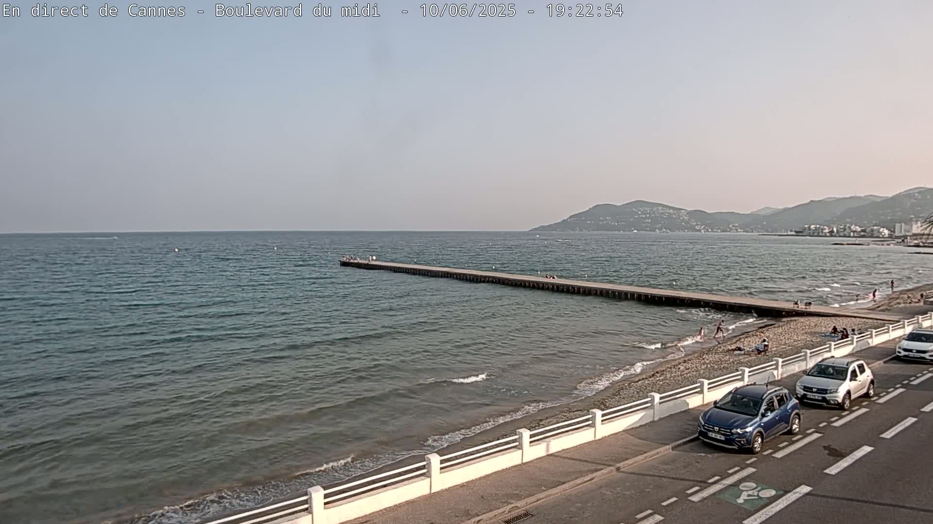 A long pier extends into a calm sea, with a sandy beach and several cars parked along a nearby road under a clear sky.