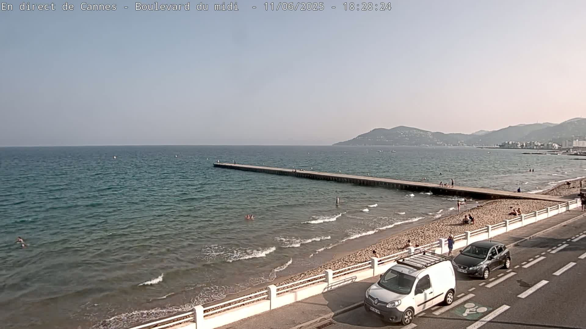 A sandy beach with calm, blue water, a long pier, and several people swimming and relaxing on a sunny day.