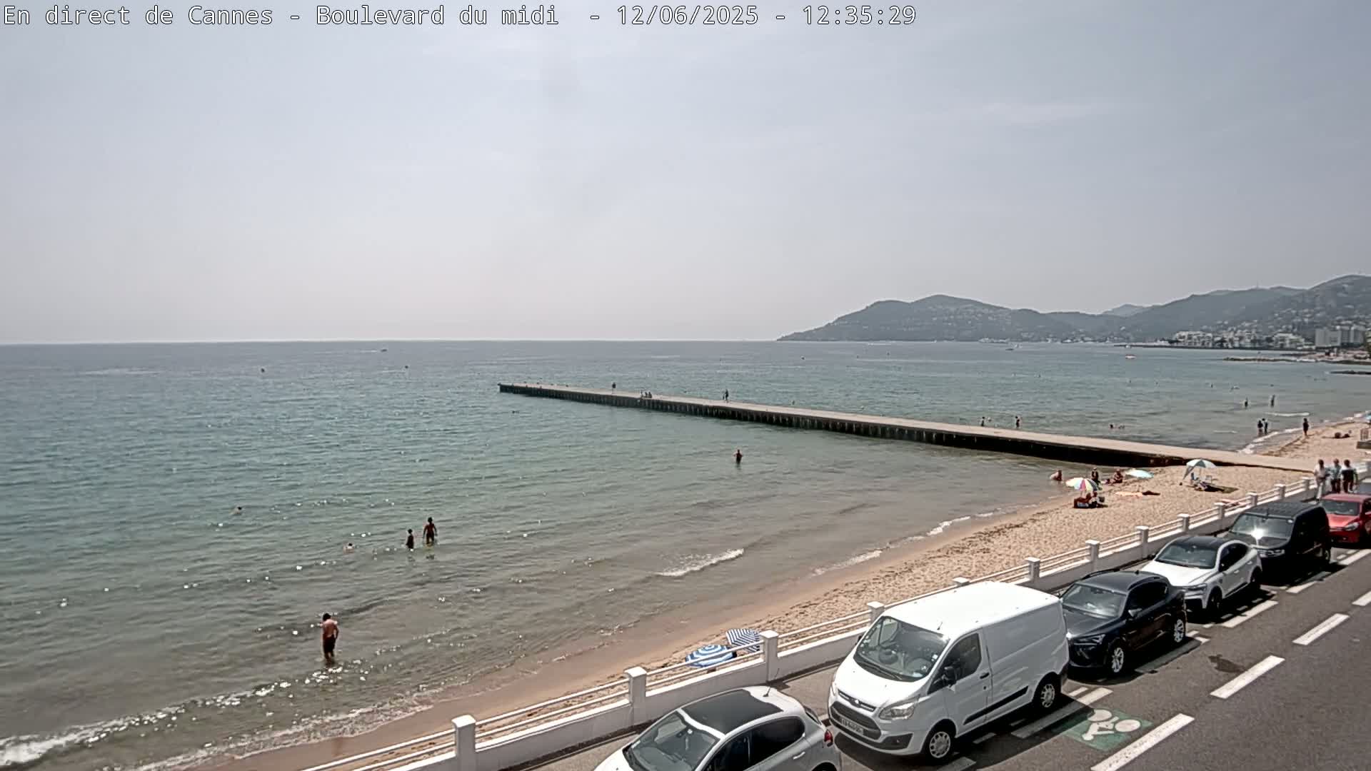 A sunny day at a beach with calm, clear water, a long pier extending into the sea, people swimming and sunbathing on the sand, and cars parked along a road next to the beach.