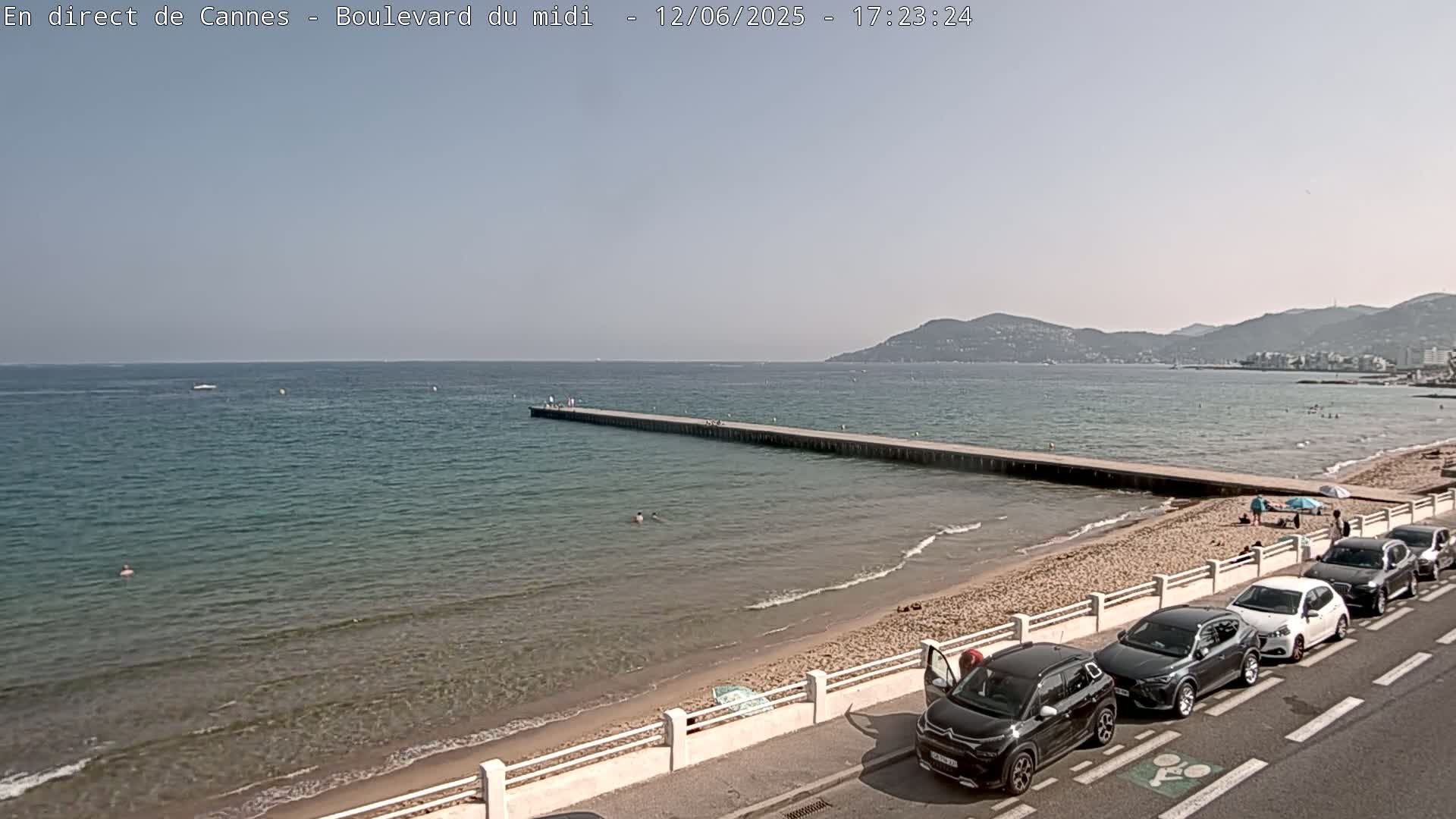 A sunny day at a beach shows a long pier extending into calm, clear water, with cars parked along the sandy shore and a few people visible on the beach and in the water.