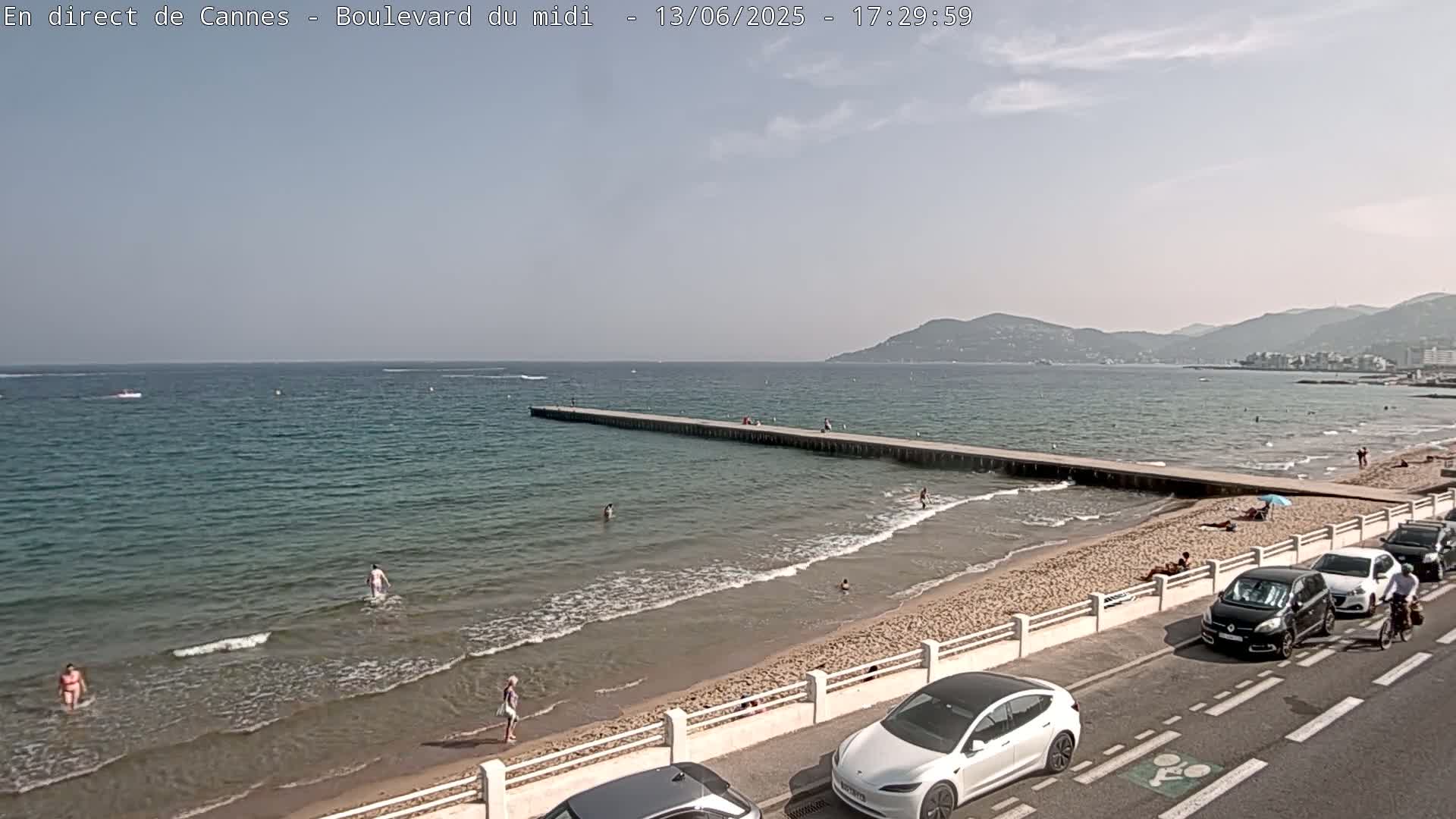 A sandy beach with calm, blue water, a long pier, people swimming and sunbathing, and cars parked along a roadside, under a partly sunny sky.
