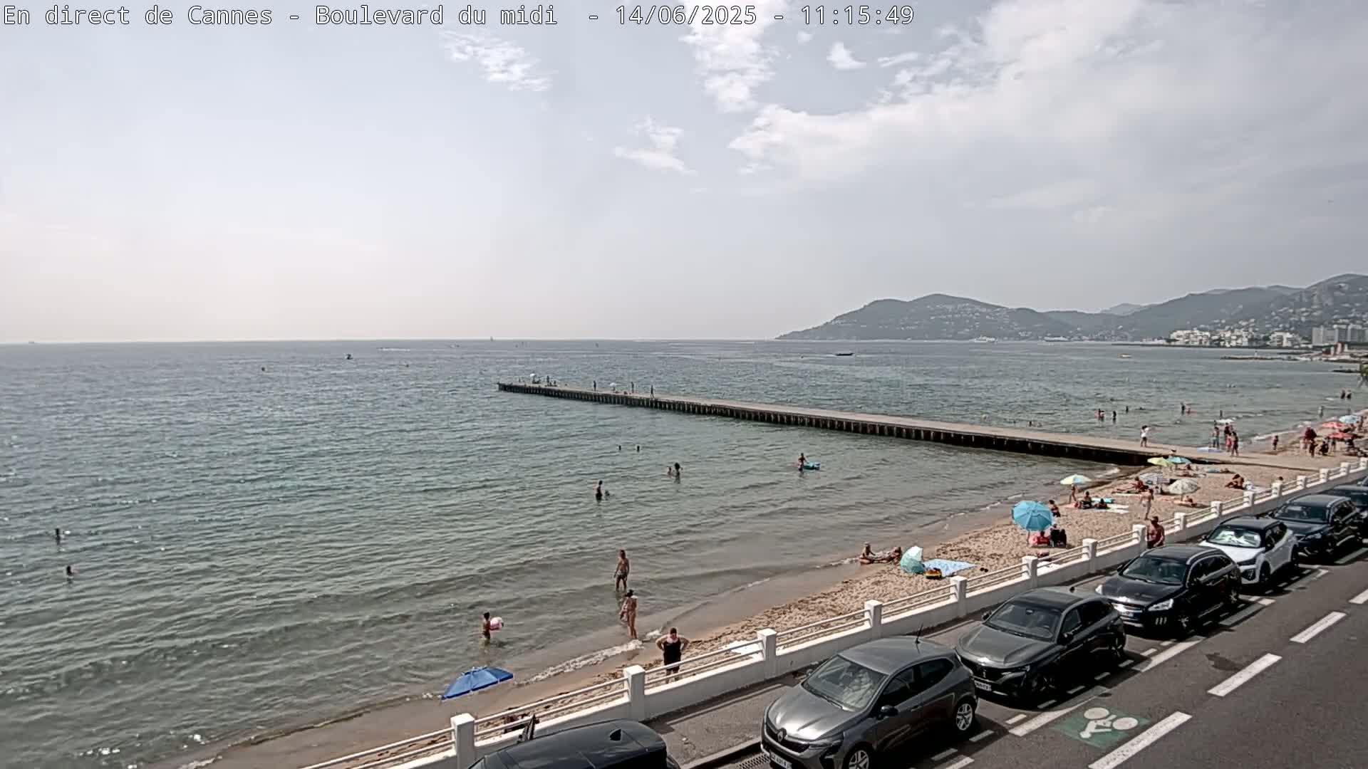 A partly cloudy day at a beach shows people swimming and sunbathing near a pier with cars parked along the roadside next to the sand.