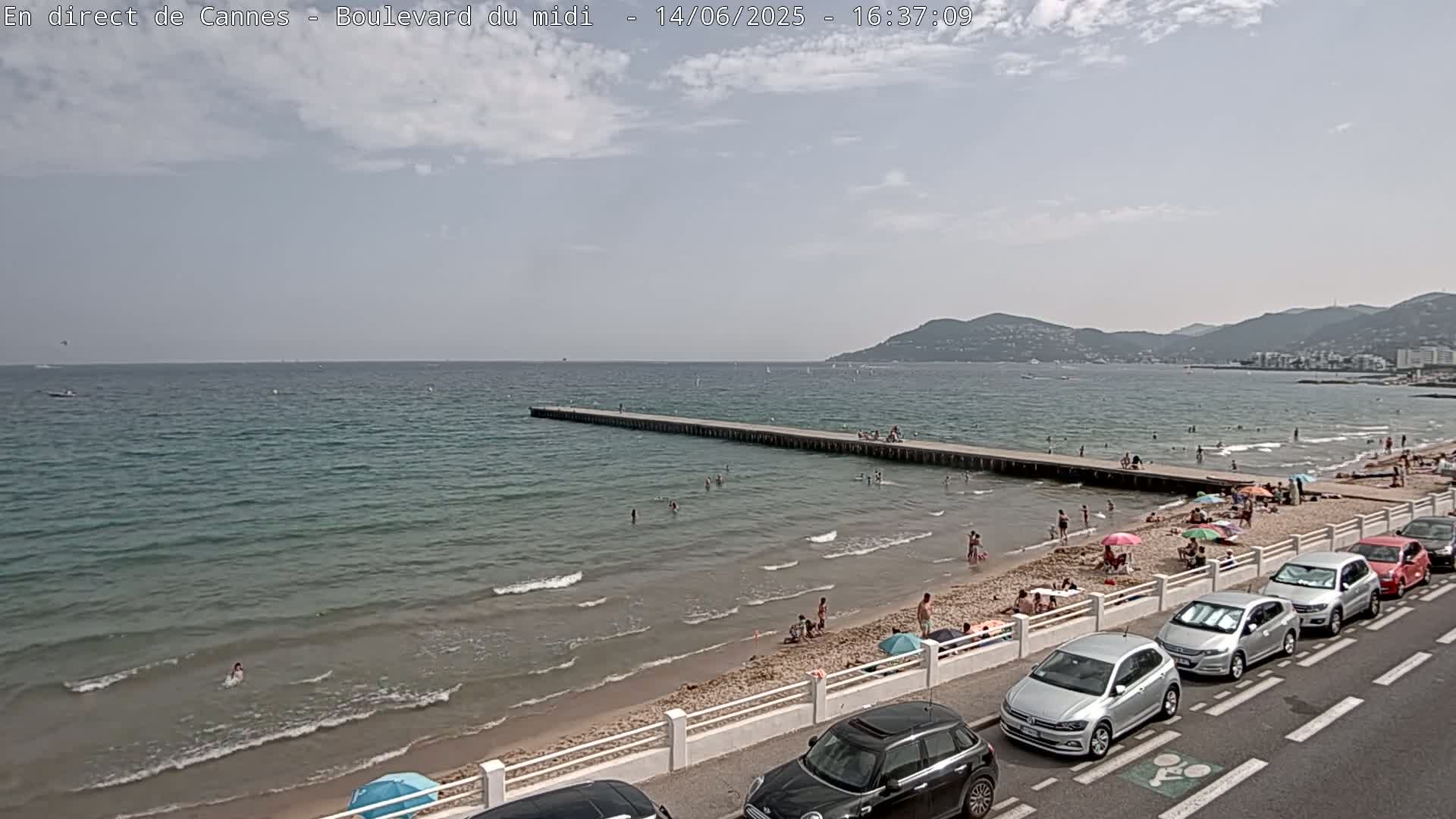 A long pier extends into a calm, light-blue sea where people are swimming and sunbathing on a sandy beach, with cars parked along the shore under a mostly cloudy sky.