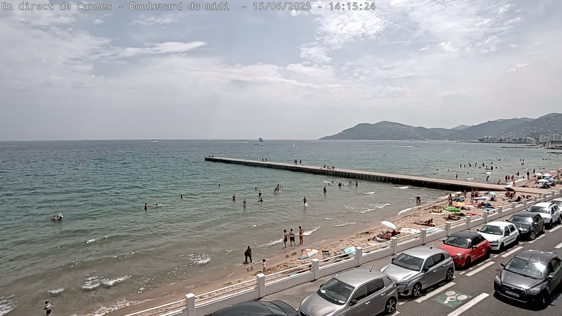 A partly cloudy day at a beach with calm, clear water, many people swimming and sunbathing, a pier extending into the sea, and cars parked along a roadway bordering the beach.