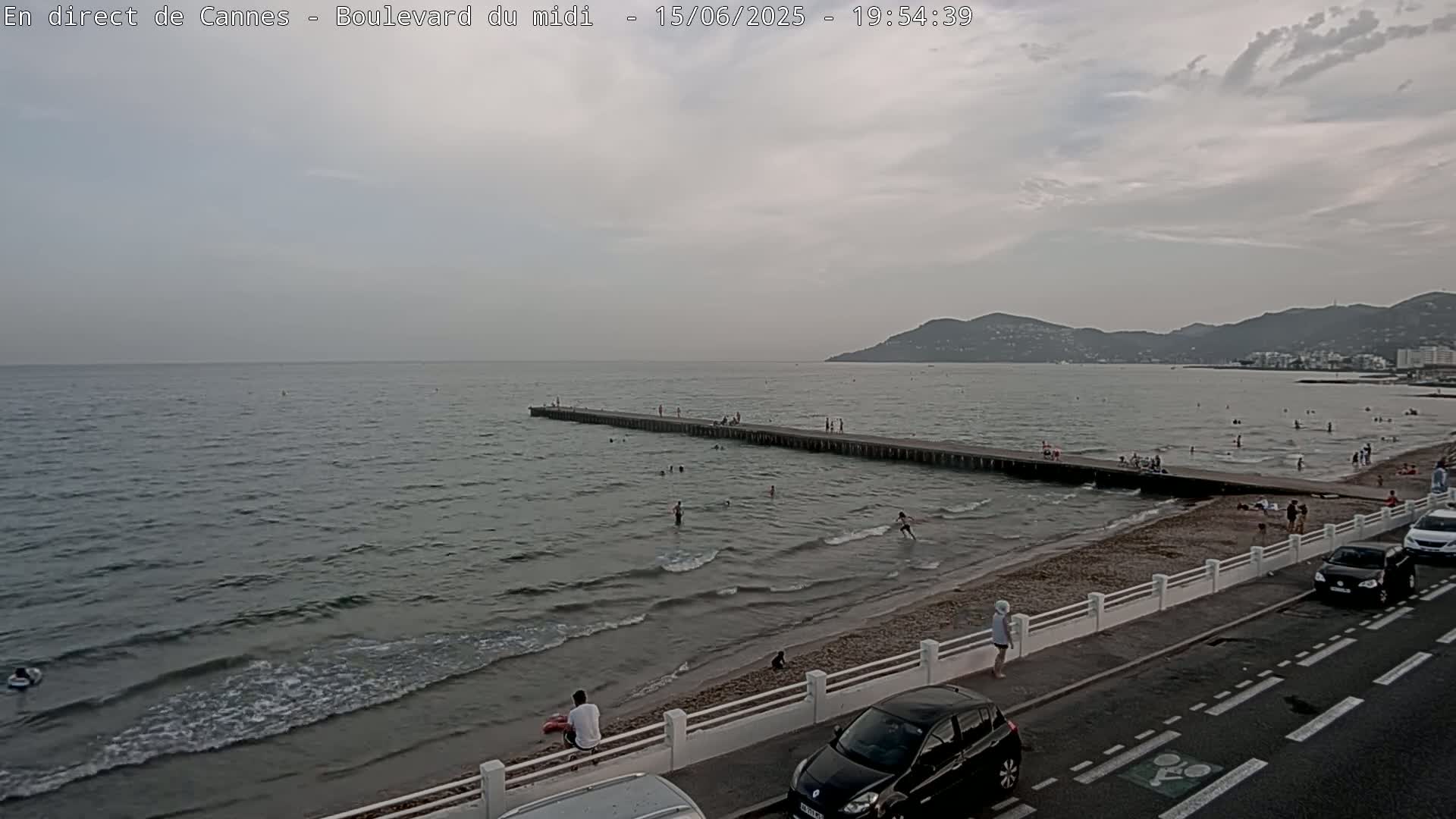 A long pier extends into a calm, gray sea, where people are swimming and sunbathing on a nearby sandy beach under an overcast sky.