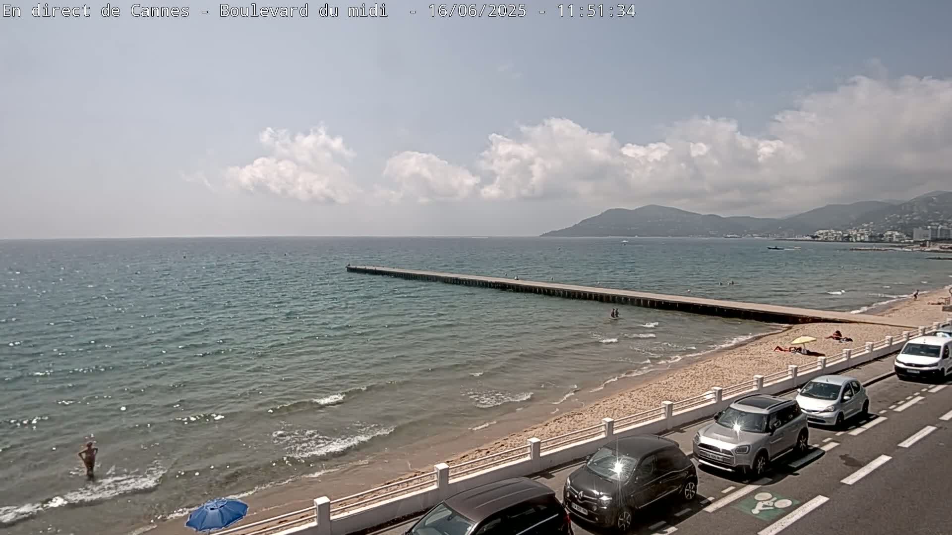 A partly cloudy day shows a long pier extending into a calm, light-blue sea from a sandy beach with a few people and several cars parked along a nearby road, and mountains visible in the distance.
