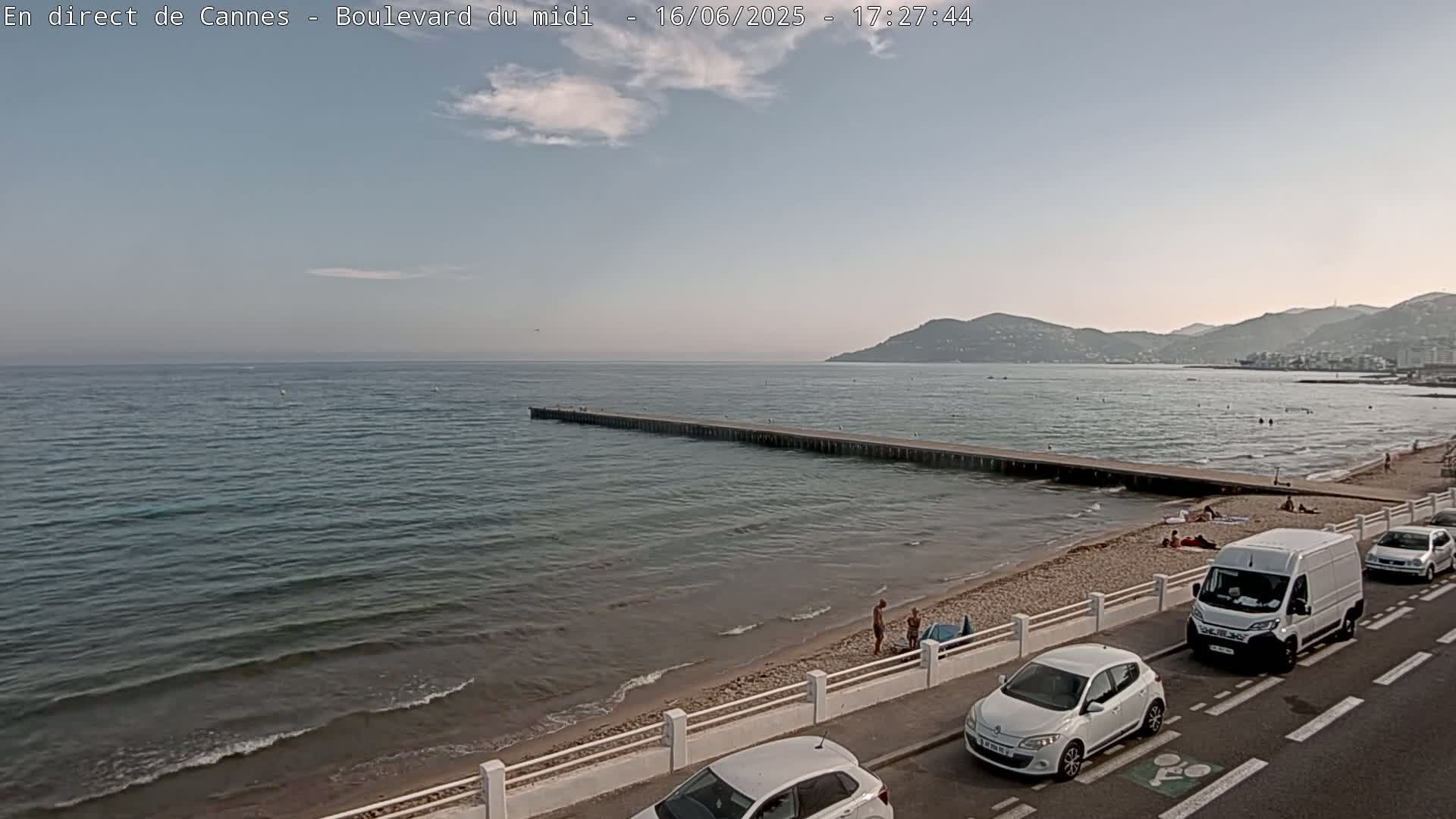 A partly sunny day shows a beach with a few people, a long pier extending into a calm sea, and cars parked along a road next to the beach, with mountains visible in the distance.