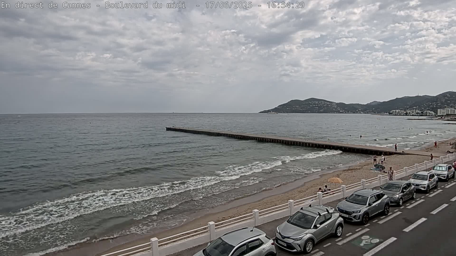 A long pier extends into a calm sea with small waves lapping a sandy beach, under a mostly cloudy sky, where several cars are parked along a roadside.