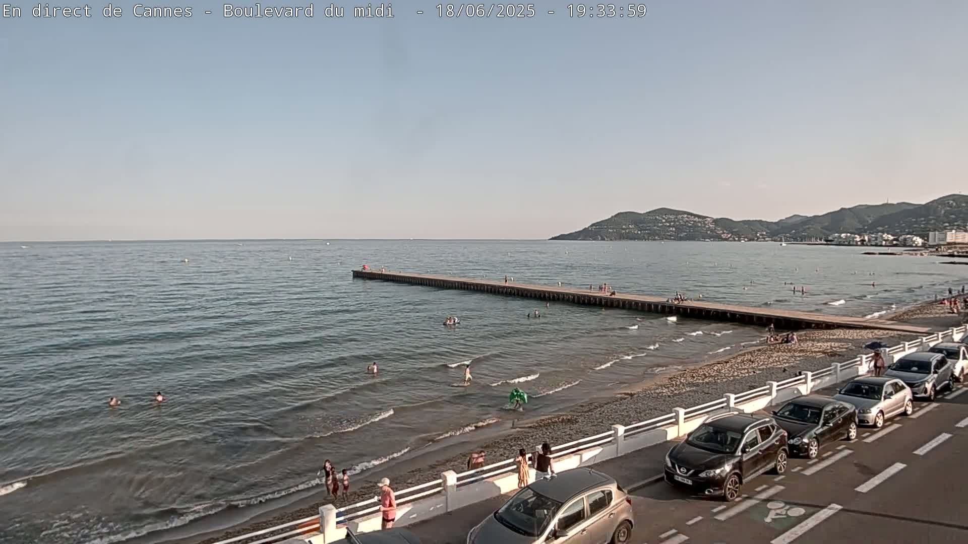 A beach scene shows a calm sea with people swimming, a long pier extending into the water, cars parked along the shore, and hills in the background under a clear sky.