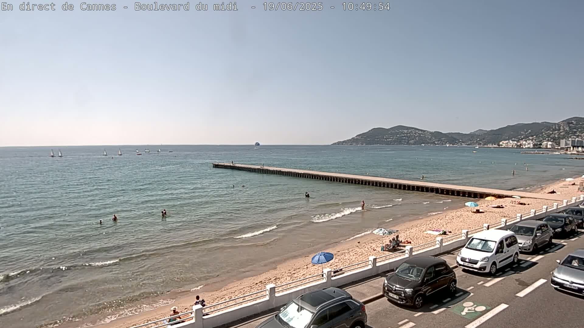 A sandy beach with people swimming and sunbathing, several cars parked along a road, a long pier extending into a calm, blue sea under a clear, sunny sky, and distant hills visible in the background.