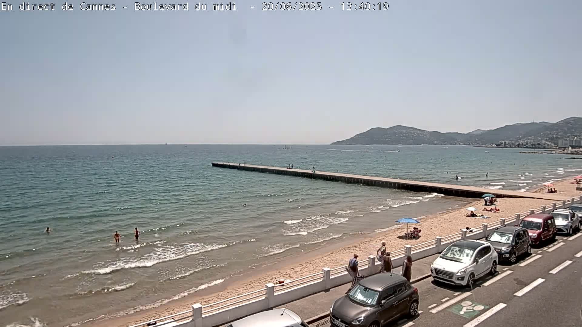 A sandy beach with calm, light blue water, a long pier, people swimming and sunbathing, and cars parked along a nearby road under a clear sky.