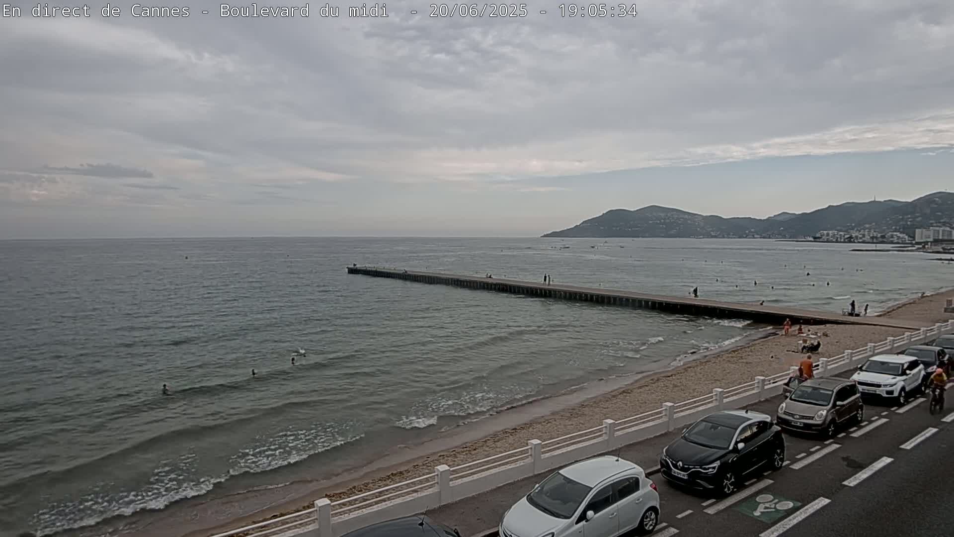 A long pier extends into a calm, gray-toned sea under an overcast sky, with a sandy beach and parked cars lining a road beyond.