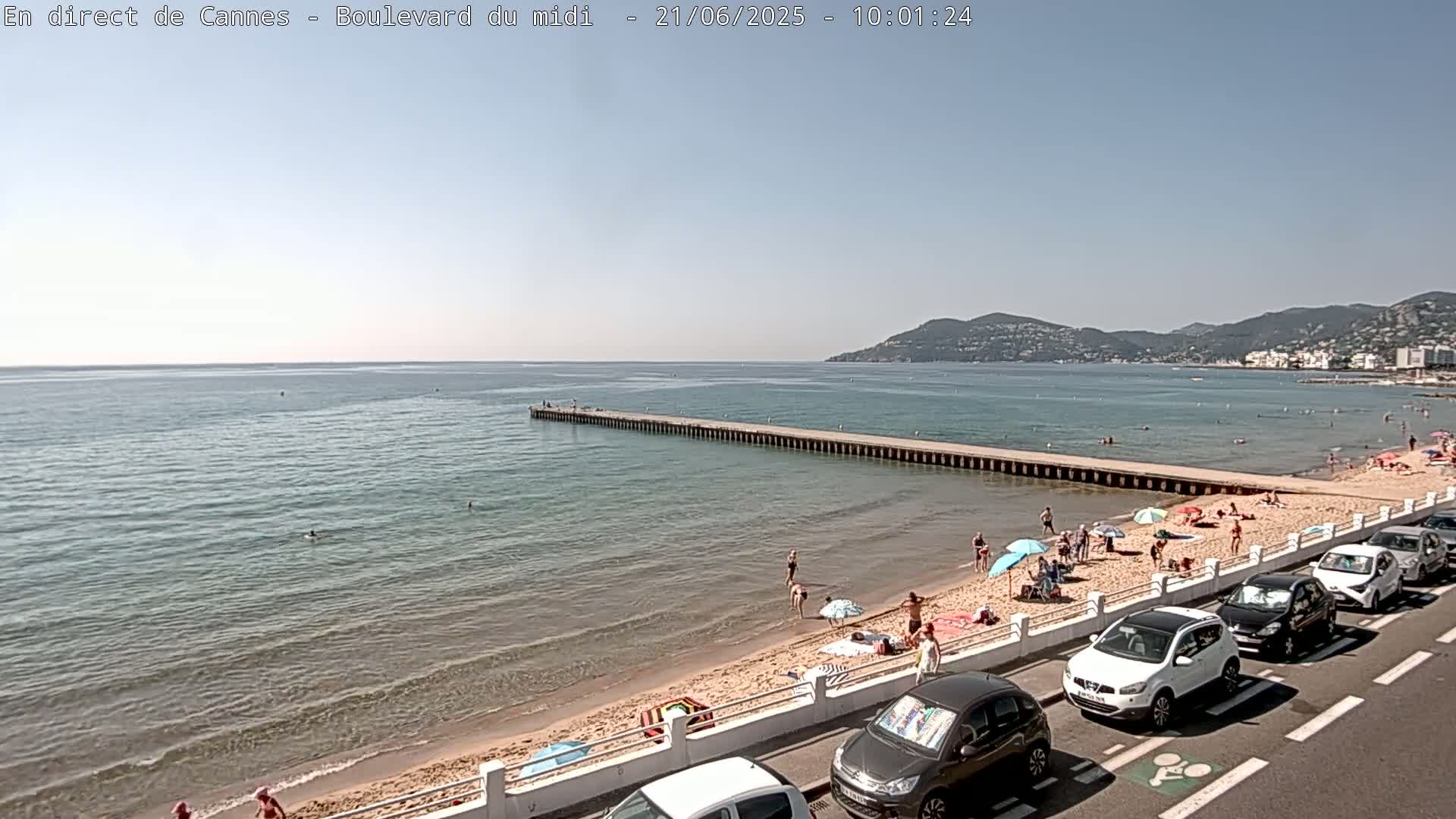 A sandy beach with people swimming and sunbathing stretches along a calm, clear ocean under a sunny sky, next to a road lined with parked cars and a long pier extending into the water.