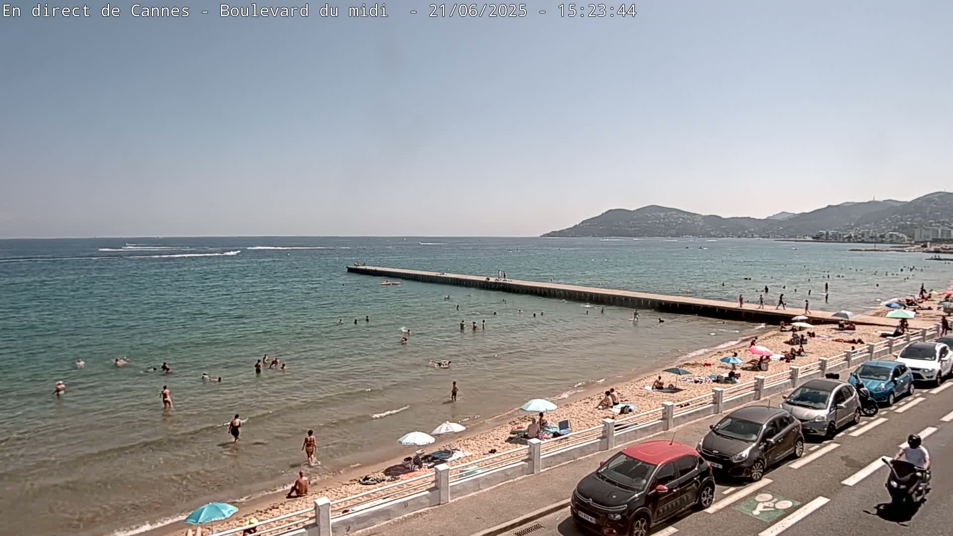 A sunny beach scene shows people swimming and sunbathing on a sandy shore next to a calm, blue sea with a long pier extending into the water; cars are parked along a road overlooking the beach.
