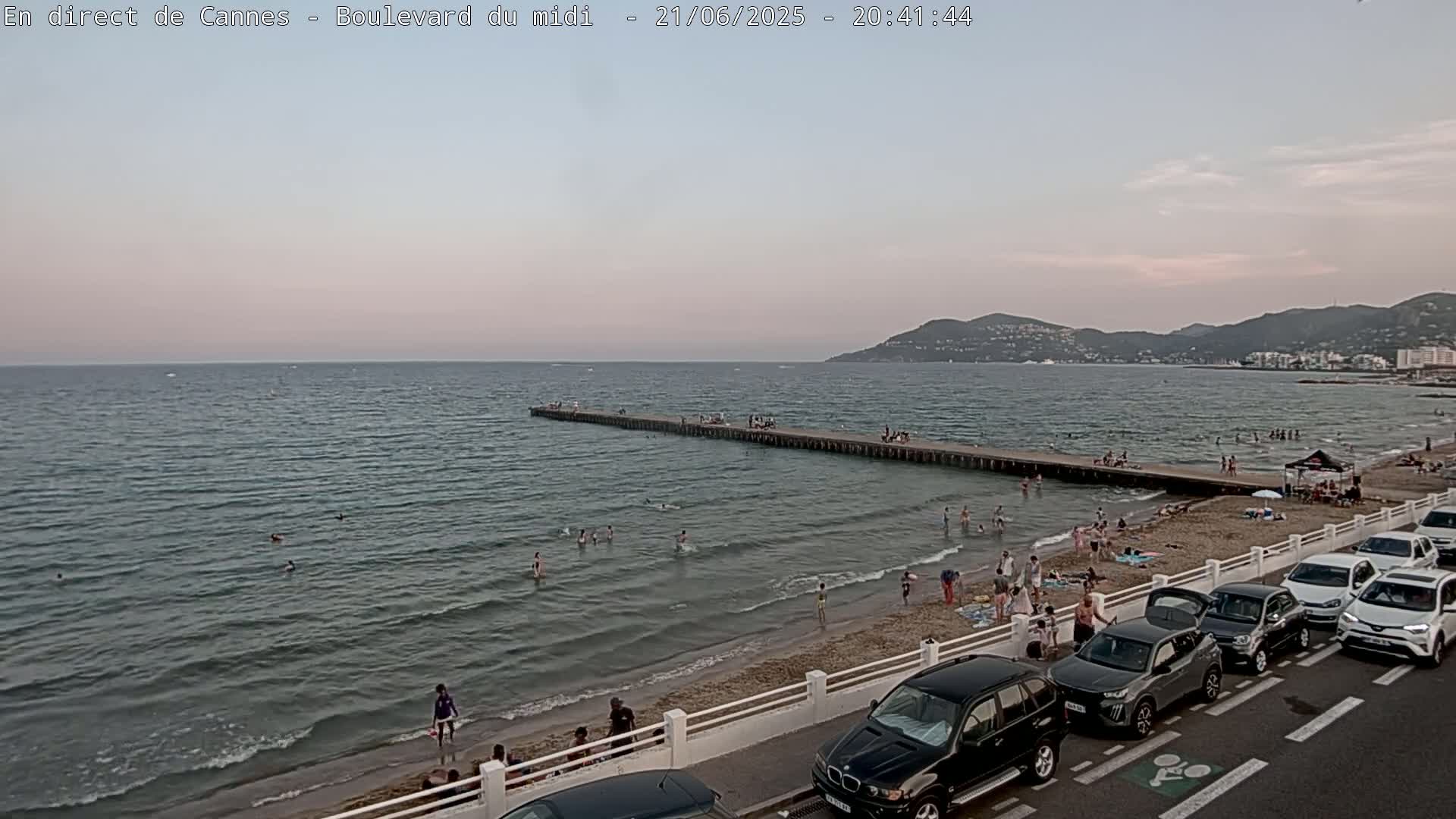 A sandy beach with people swimming and sunbathing, a pier extending into a calm sea under a twilight sky, and cars parked along a road next to the beach.