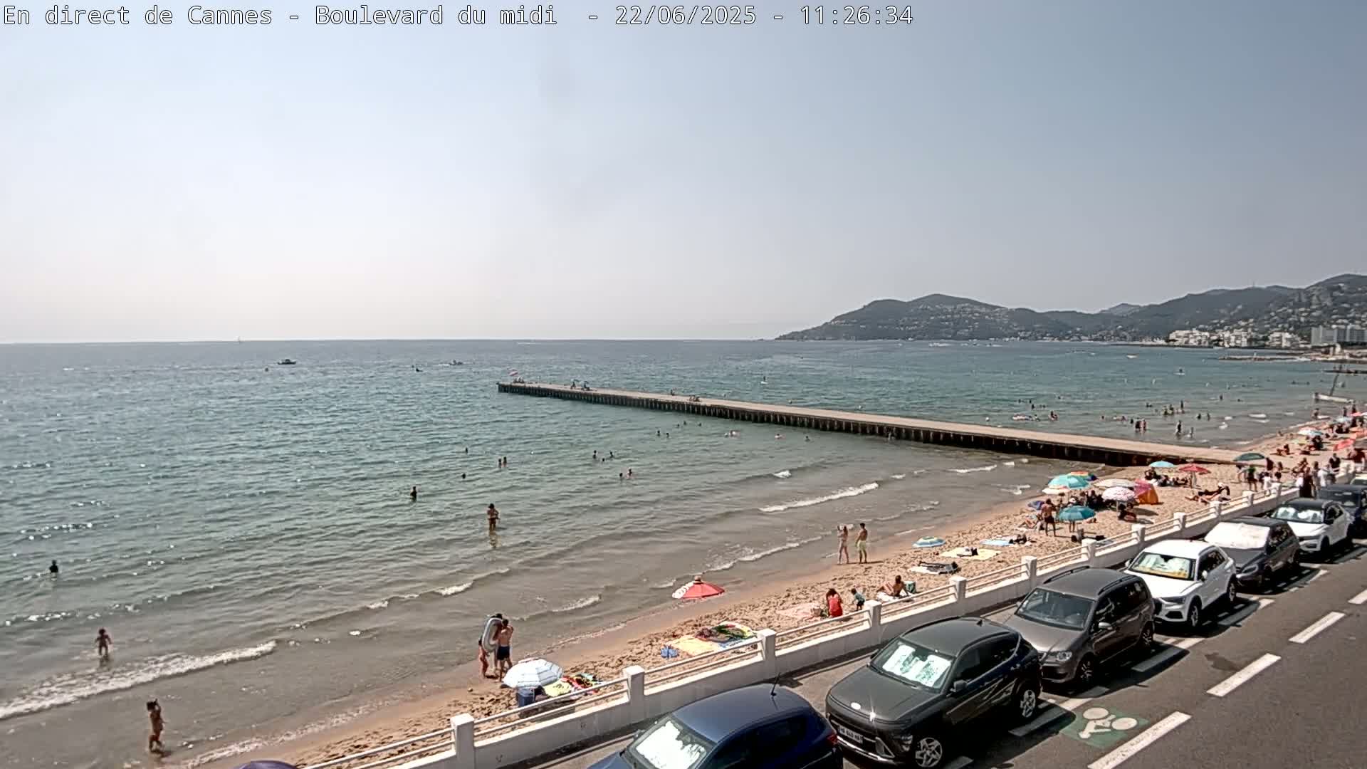 A long pier extends into a calm, light-blue ocean where people swim, while a sandy beach is crowded with sunbathers and parked cars line the road.