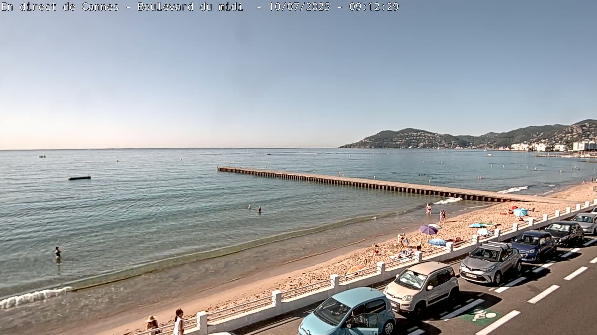 A sandy beach with calm, clear water, several people swimming and sunbathing, cars parked along the roadside, and a pier extending into the sea under a clear, sunny sky.