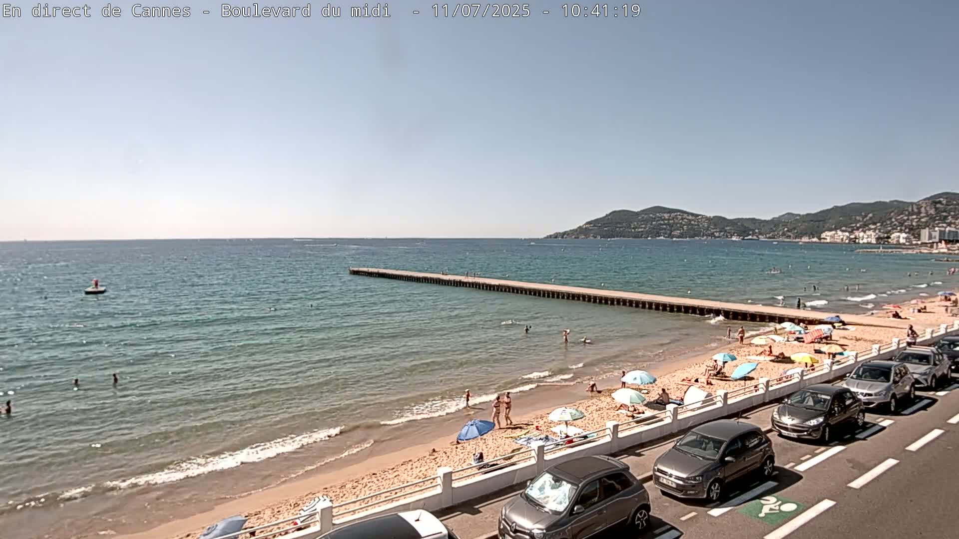 A sandy beach with people sunbathing under umbrellas, parked cars along a road, and a long pier extending into a calm, blue sea under a clear sky.