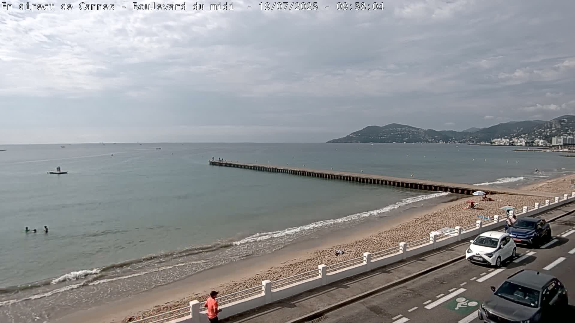 A mostly cloudy day shows a sandy beach with a long pier extending into a calm sea, a few people on the beach and in the water, and cars parked along a roadside.