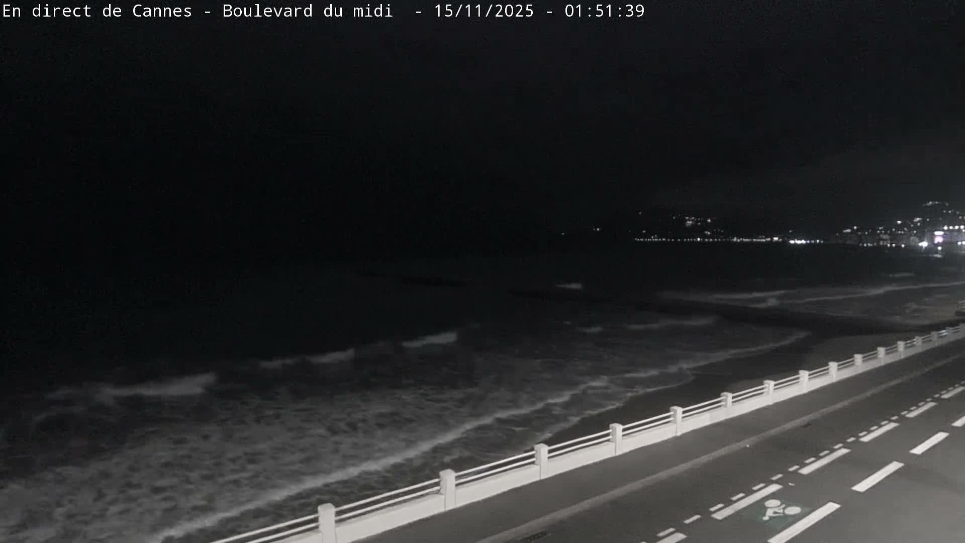 The image displays a dark night view of a coastal area with moderate waves along the shore, a road and promenade in the foreground, and distant city lights illuminating the horizon under clear weather conditions.