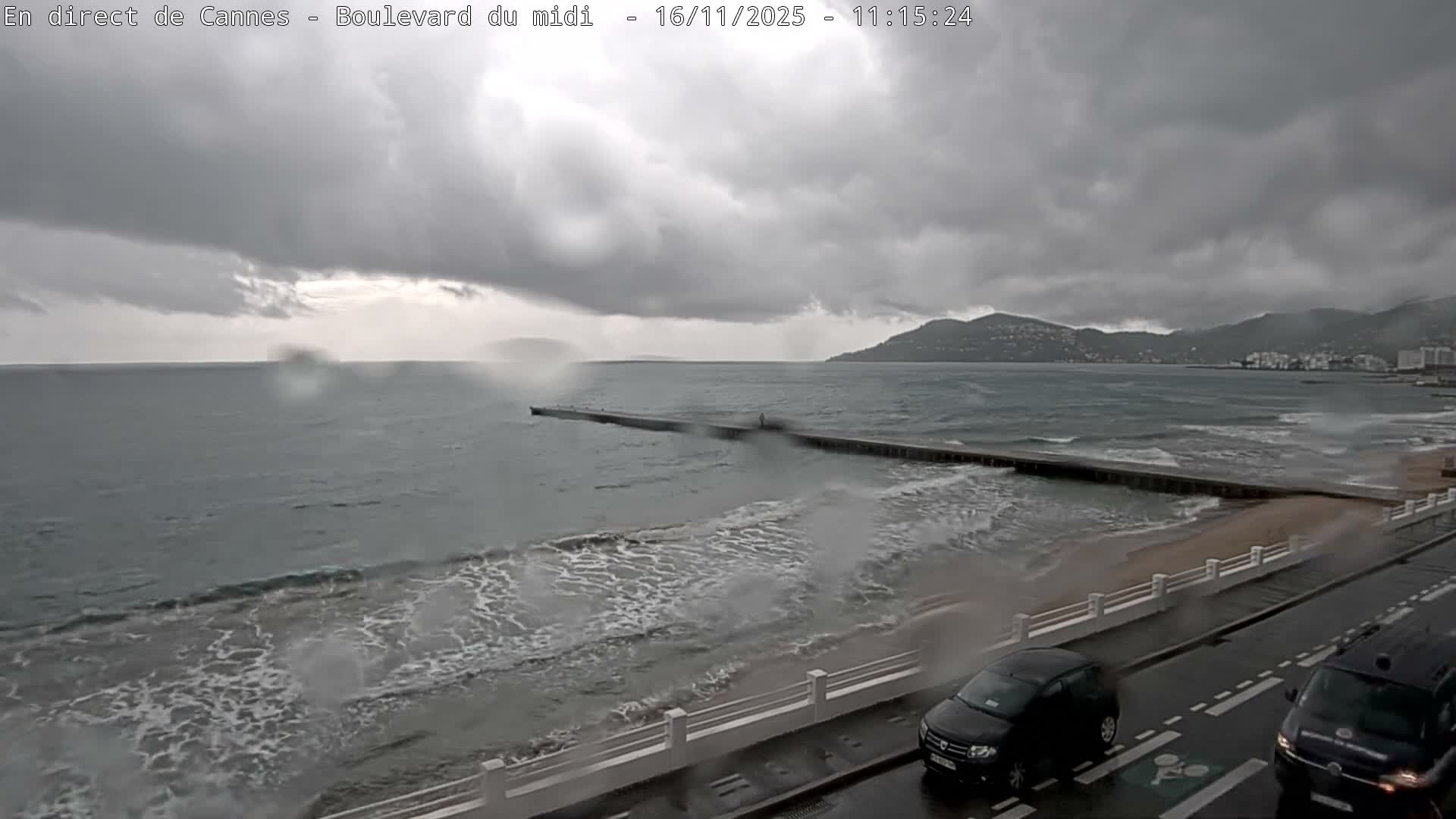 On an overcast and likely rainy day, strong waves crash against a long pier and a wet sandy beach, with distant mountains shrouded in clouds and two cars traveling on a wet coastal road.