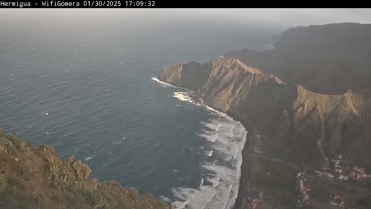 A high-angle view shows a rocky coastline with crashing waves on a somewhat hazy day.