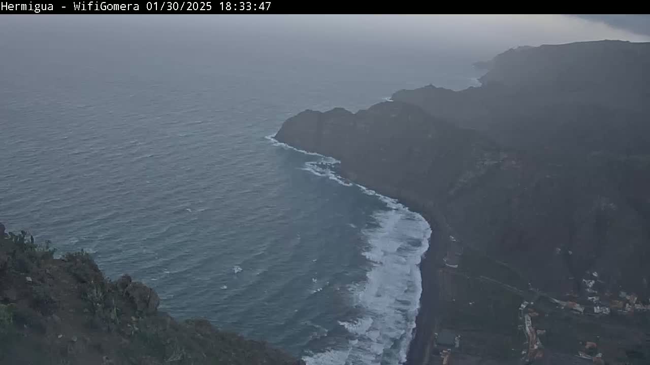 A hazy, overcast view shows a dark, rocky coastline meeting a dark-blue ocean with white-capped waves.