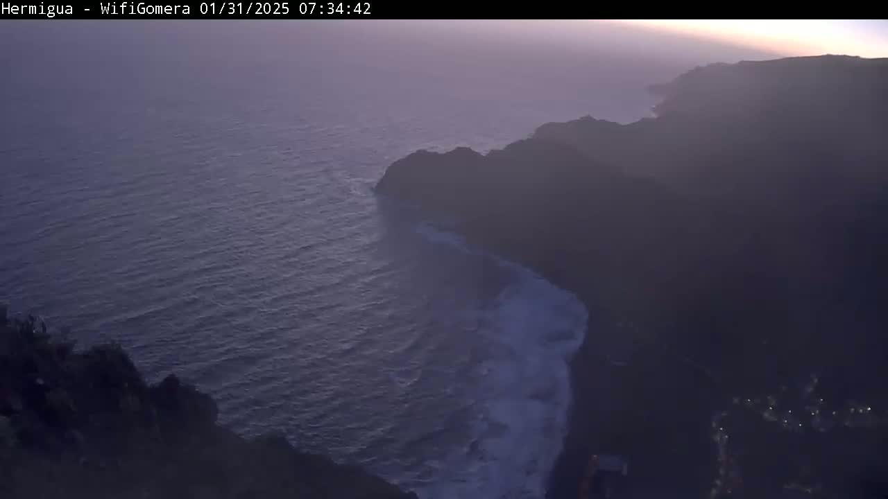 A hazy, dusky view shows dark, rocky cliffs meeting a dark blue ocean with white waves crashing along the shore.
