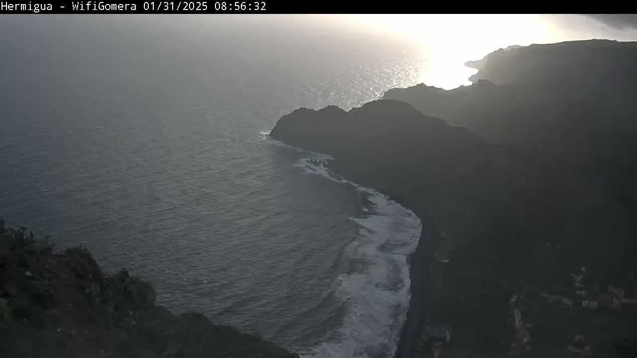 A hazy sunrise illuminates a dark, rocky coastline where waves crash on a black sand beach.