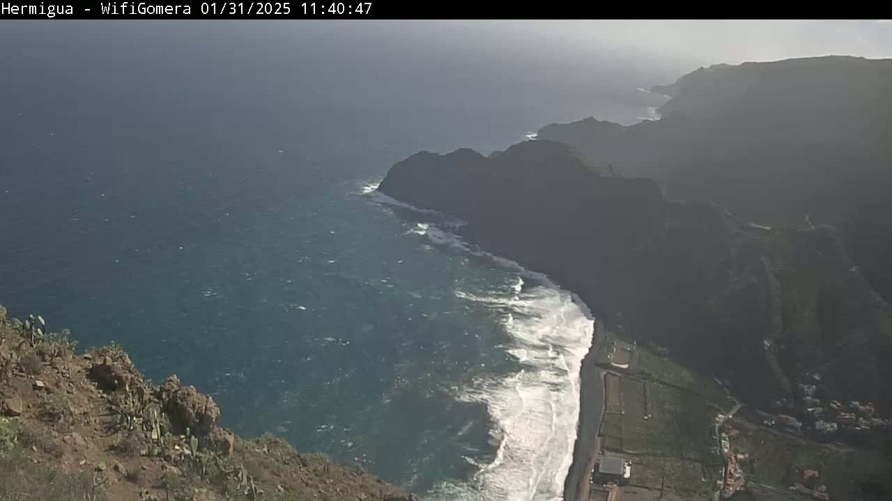 A high-angle view shows a rocky coastline with a beach where waves crash against the shore, under hazy, sunny conditions.