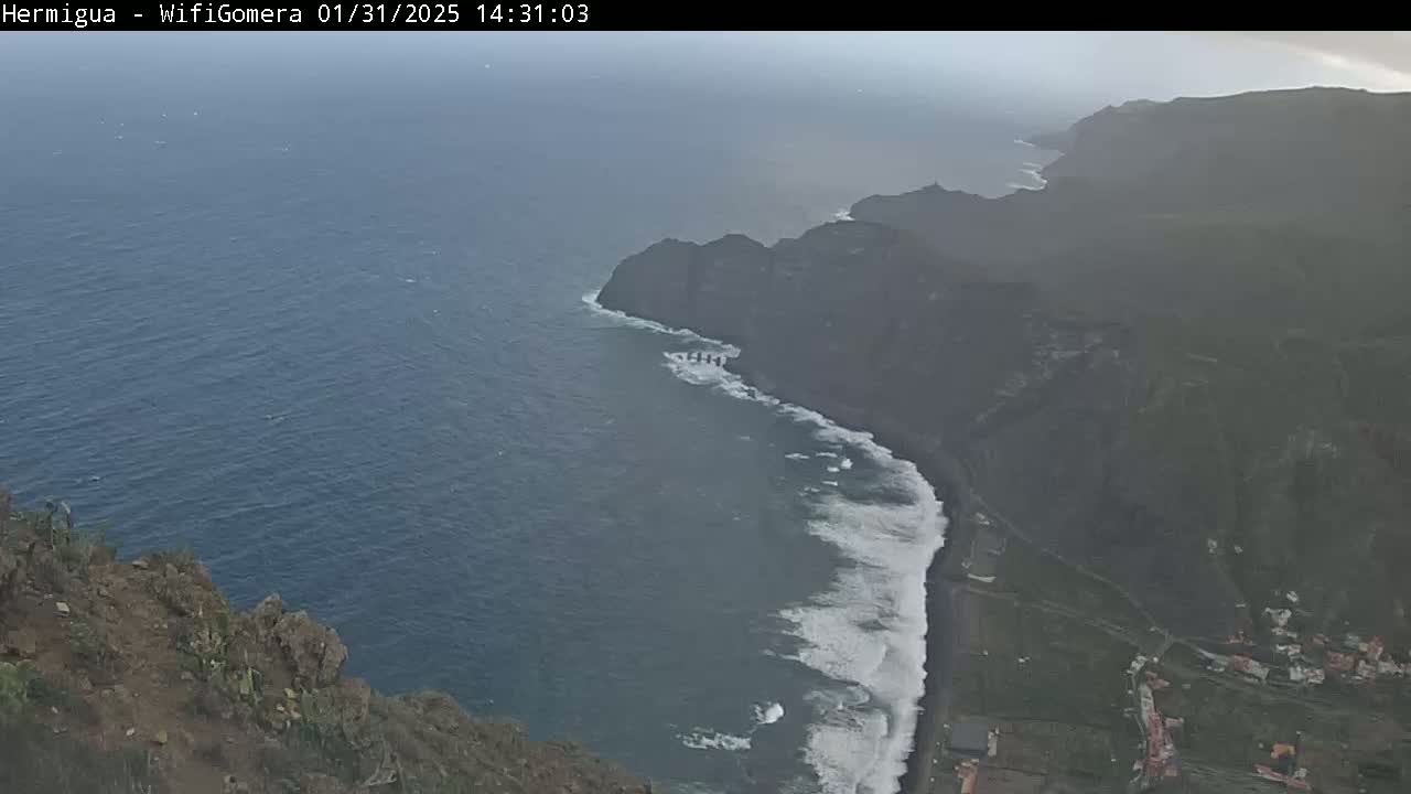 A hazy coastal view shows dark cliffs meeting a dark-blue ocean with white-capped waves crashing on a black sand beach.