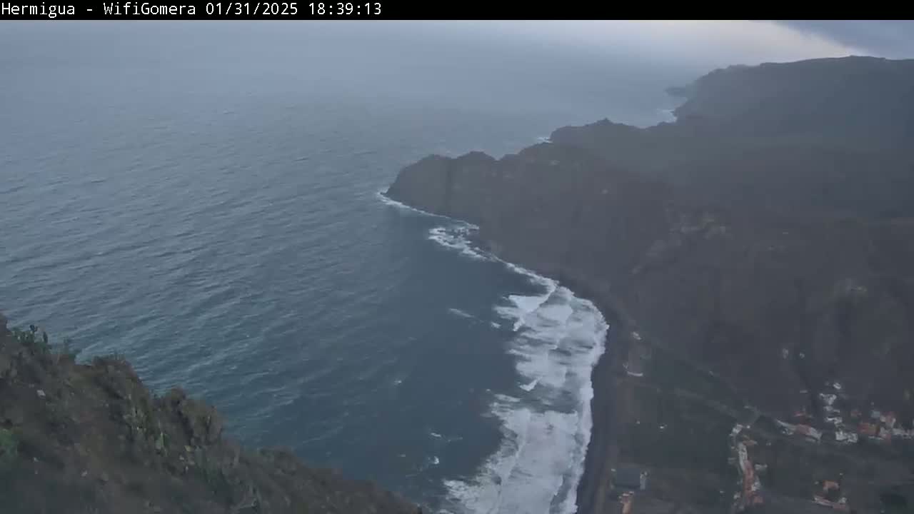 A dark, rocky coastline with a dark-sand beach and crashing waves is seen from an elevated viewpoint on an overcast day.