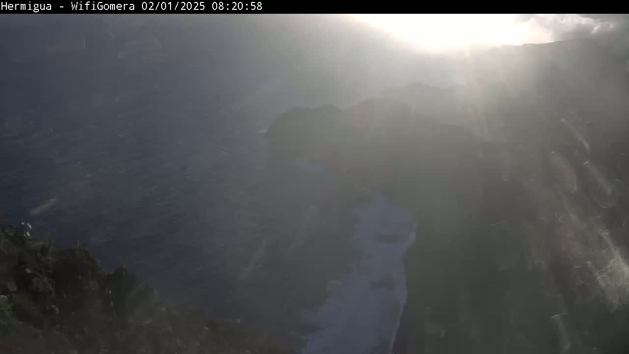 A hazy, sunlit view from a high vantage point shows dark-colored ocean water and a rocky, dark-colored coastline.