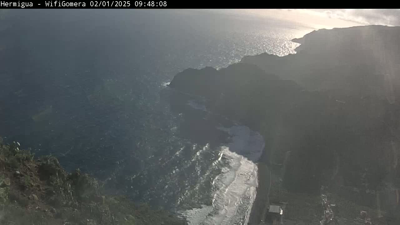 A high-angle view shows a rocky coastline meeting a dark, wavy ocean under a partly sunny sky.