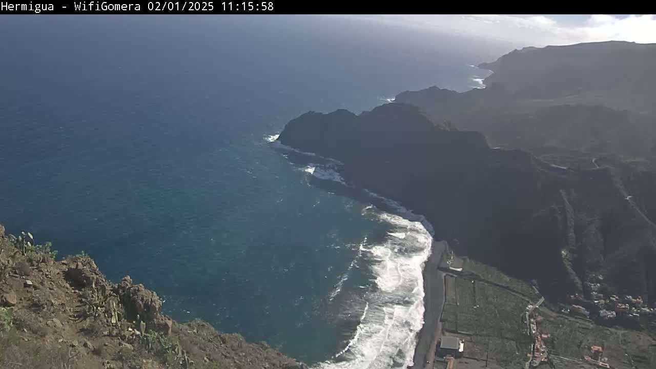 A high-angle view shows a dark-colored, rocky coastline with a black-sand beach and crashing waves under a partly sunny sky.
