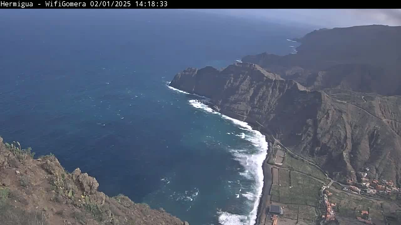 A high-angle view shows a rocky, dark-colored coastline meeting a dark-blue ocean with white-capped waves under a mostly clear sky.