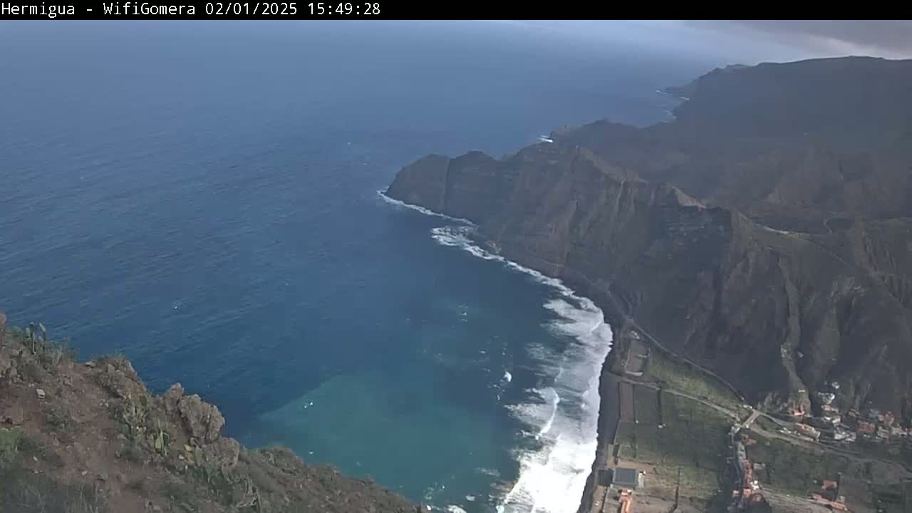 A high-angle view shows a dark, rocky coastline meeting a deep blue ocean with white waves crashing against the shore under partly cloudy skies.