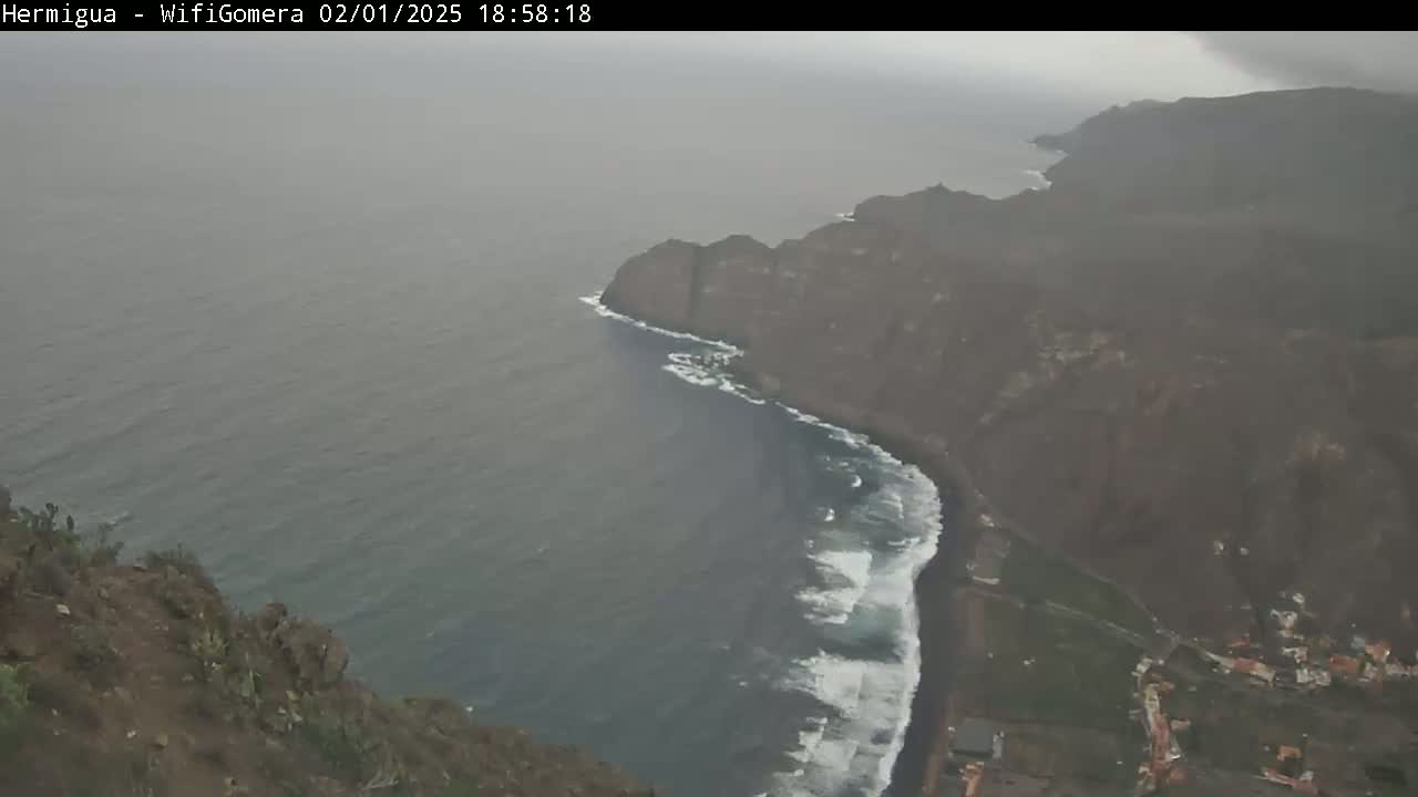 A high-angle, hazy view shows a dark-colored coastline with waves crashing on a beach below a steep, rocky cliff during overcast weather conditions.