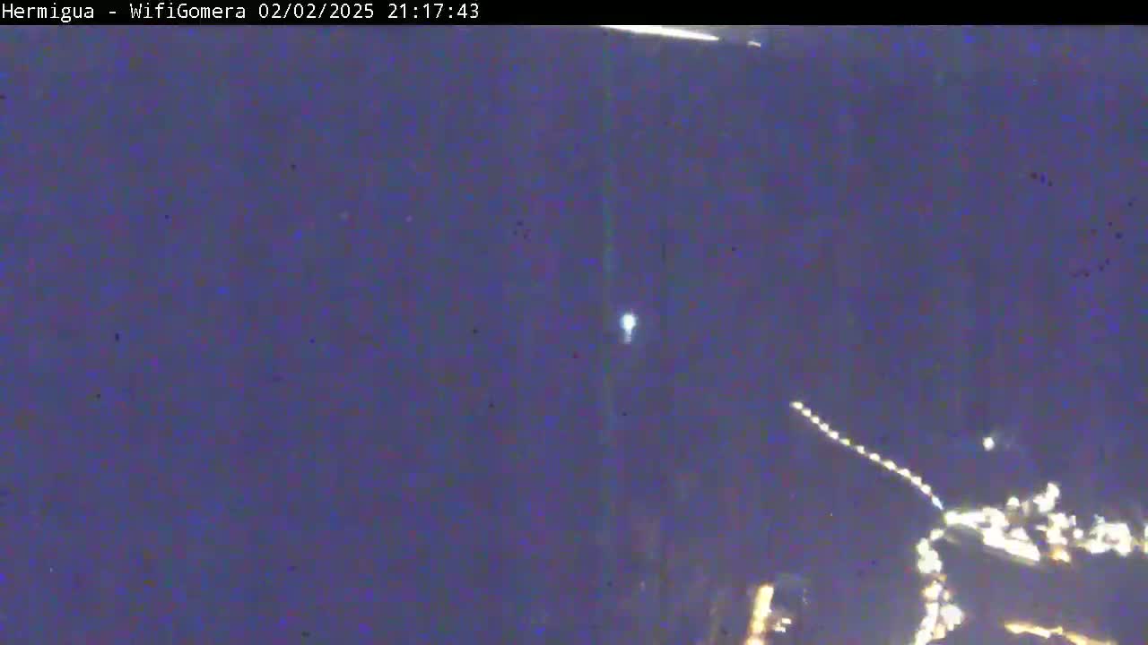 A nighttime, low-light image shows a blurry view of what appears to be a city or town lights at the bottom of the frame, with a bright light and what appears to be a streak of light in the night sky above.