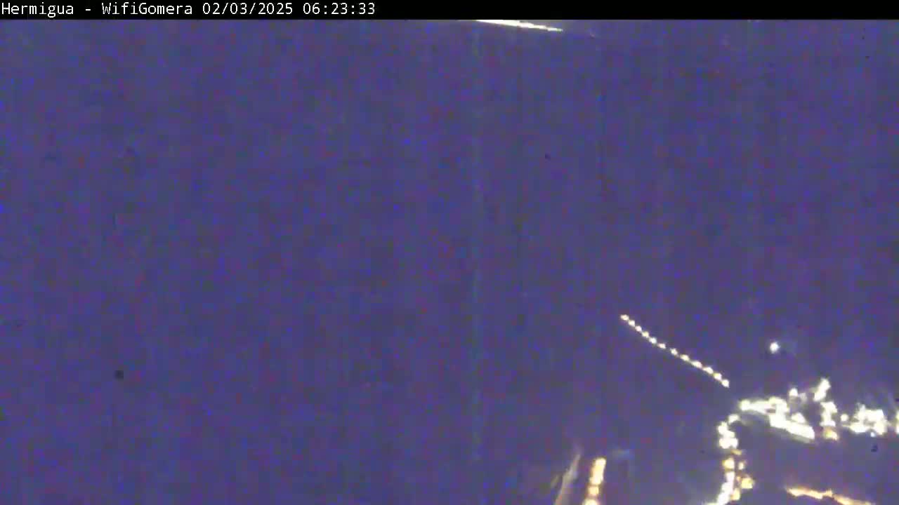 A low-light nighttime image shows a blurry view of lights possibly from a town or city in the distance, with a faint, possibly streaking light source in the sky.