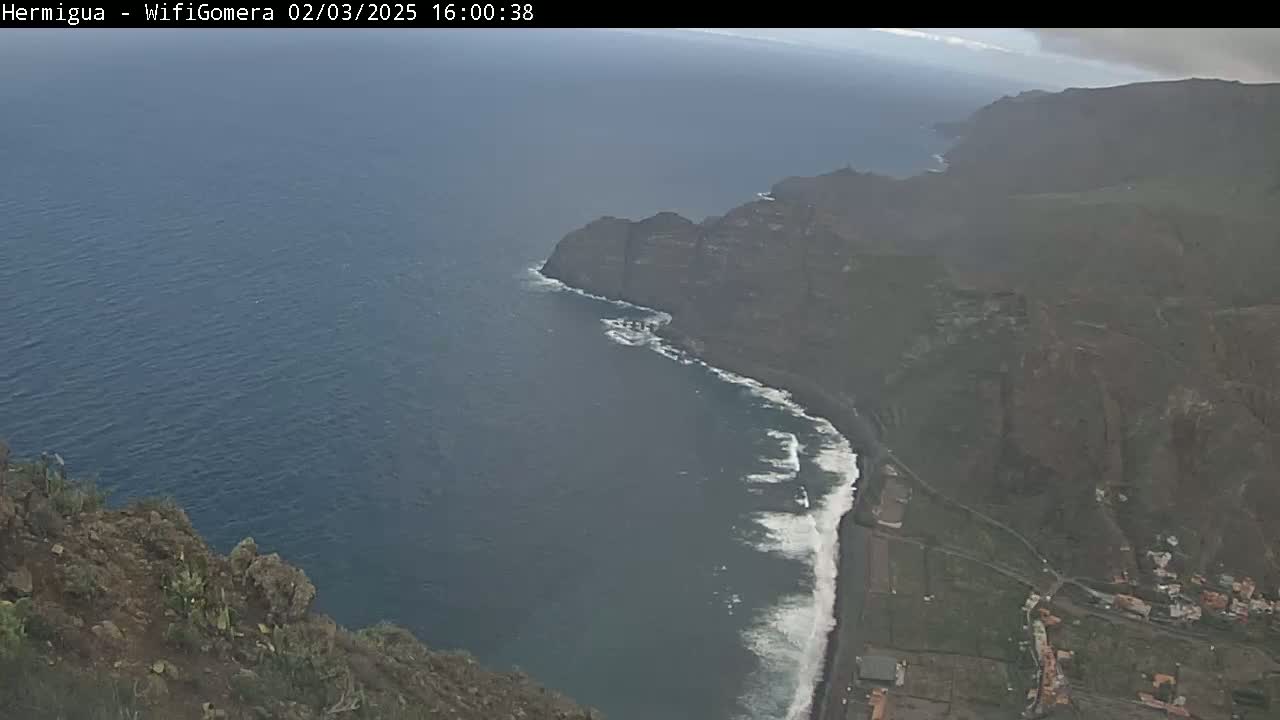 A high-angle view shows dark-colored cliffs meeting a dark-blue ocean with white waves along the shore, under somewhat cloudy skies.