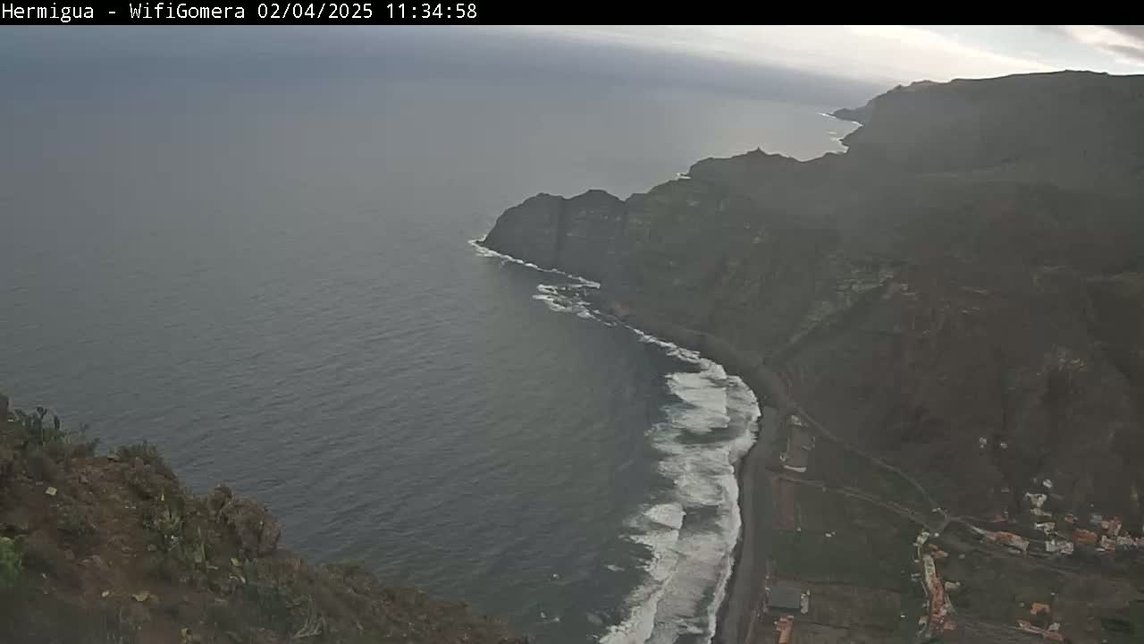 A high-angle view shows a dark, rocky coastline with crashing waves on a partly cloudy day.