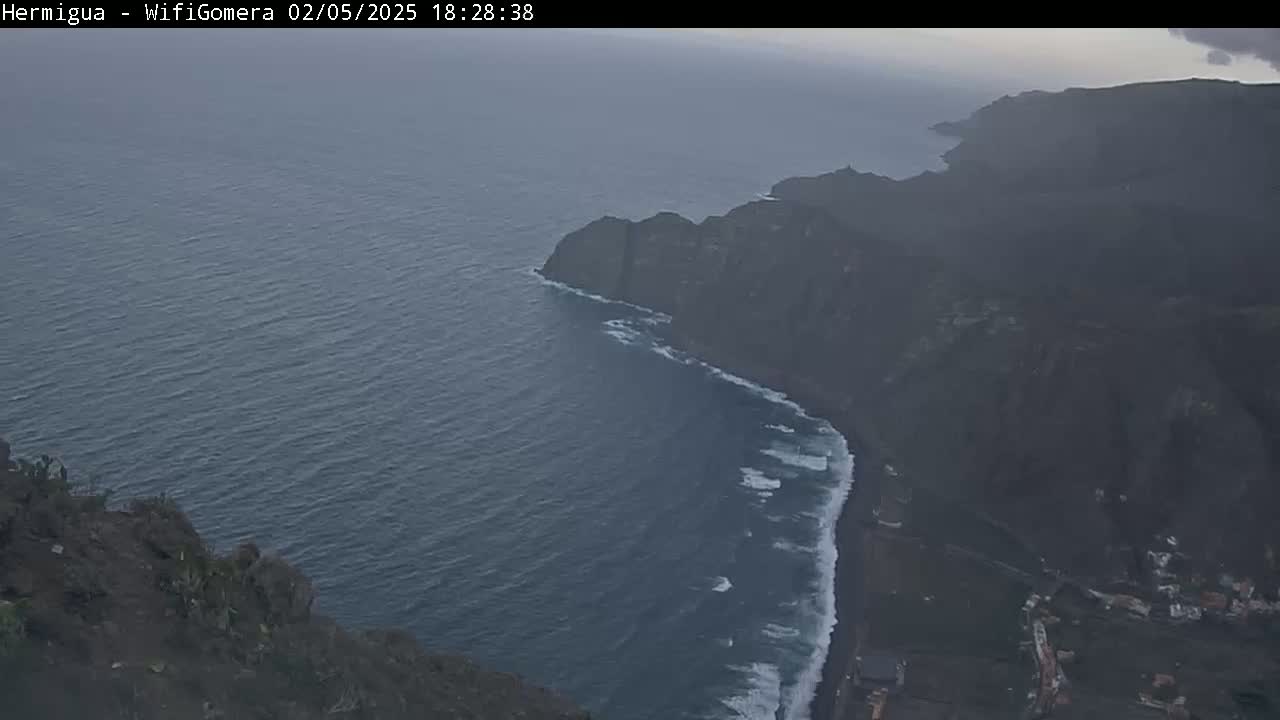 A high-angle, long shot shows dark volcanic rock cliffs meeting a dark blue ocean with small waves crashing on the shore under an overcast sky.