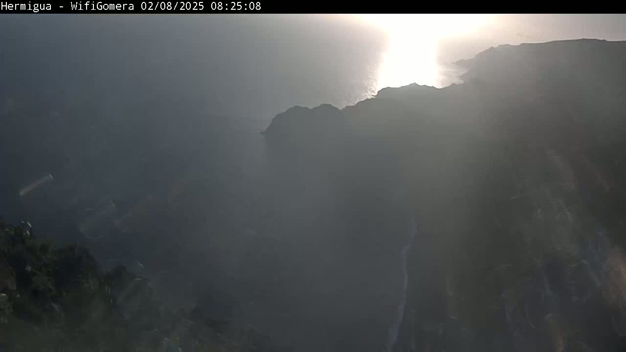 A hazy sunrise illuminates a dark, rocky coastline overlooking a body of water.